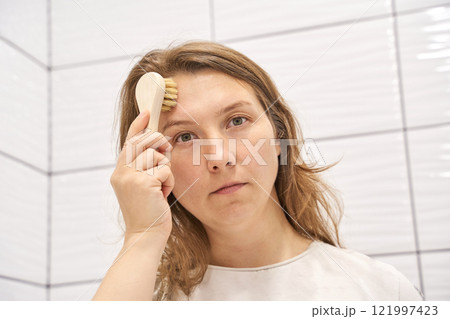 Young caucasian female using facial brush for skincare routine in bathroom Young caucasian female using facial brush for skincare routine in bathroom 121997423