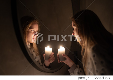 A young woman gazes into a mirror while holding a lit candle, the warm glow illuminating her face and its reflection. The serene moment evokes introspection and quiet elegance 122001705