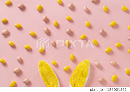 Easter concept with yellow bunny ears, pastel chocolate candy sweets as eggs on pink background. View from above. Flat lay minimal style. Happy Easter greeting card. 122001831