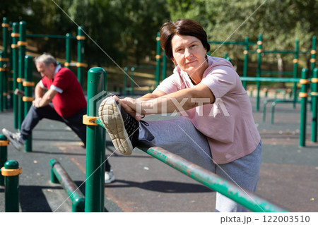 Positive elderly woman doing leg stretching on outdoor sports ground 122003510