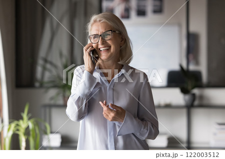 Mature businesswoman wear glasses standing indoors while talking on smartphone Mature businesswoman wear glasses standing indoors while talking on smartphone 122003512