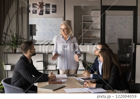 Businesswoman standing and addressing group of professionals seated at table 122004059