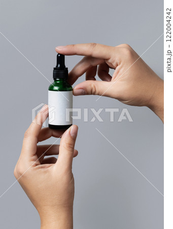 Hands holding green dropper bottle with blank label against grey close up, cosmetic mockup 122004242