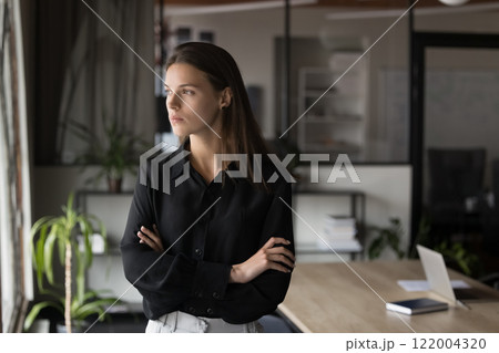 Young woman standing near window, gazing outside with contemplative expression 122004320