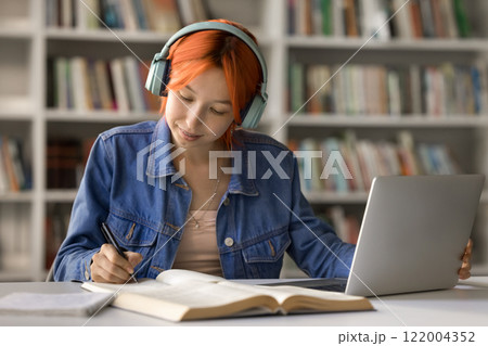 Young female student in headset sitting by notebook taking notes 122004352