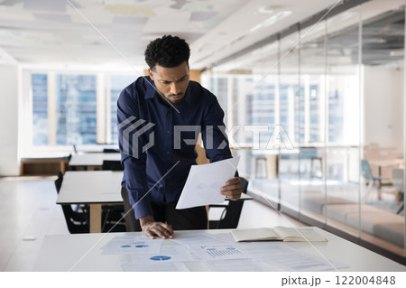Focused concerned young African professional man reviewing marketing report Focused concerned young African professional man reviewing marketing report 122004848