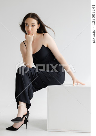 Fashion model posing sitting on white cube wearing black dress and high heels 122005241
