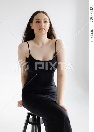 Fashion model posing wearing a tight black dress sitting on a stool 122005320
