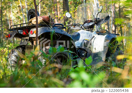Quad bike or ATV in the tall grass in sunny forest, side view. An all terrain vehicle rests in a dense forest, surrounded by tall trees and thick underbrush Quad bike or ATV in the tall grass in sunny forest, side view. An all terrain vehicle rests in a dense forest, surrounded by tall trees and thick underbrush 122005436