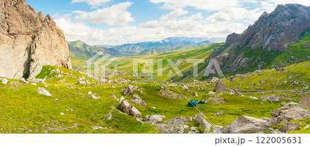 In the image, a green tent is set up at the foot of the cliffs under a blue sky with white clouds. Green grass and many stones typical of the nature of Kyrgyzstan are spread around In the image, a green tent is set up at the foot of the cliffs under a blue sky with white clouds. Green grass and many stones typical of the nature of Kyrgyzstan are spread around 122005631