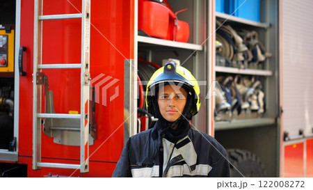 Portrait of confident female firefighter against the background of big red truck. Young firewoman in protective uniform looking at camera near a fire engine. Saving lives and heroic profession concept Portrait of confident female firefighter against the background of big red truck. Young firewoman in protective uniform looking at camera near a fire engine. Saving lives and heroic profession concept 122008272