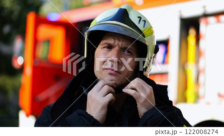 Young fireguard in protective uniform putting helmet against background of a big red truck. Male firefighter in full equipment near fire engine ready to work. Saving lives and heroic profession Young fireguard in protective uniform putting helmet against background of a big red truck. Male firefighter in full equipment near fire engine ready to work. Saving lives and heroic profession 122008274