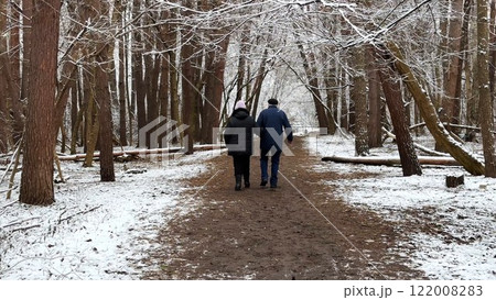 Mature couple going on snowy path at winter pine forest. Senior husband and wife walking through trail at coniferous woodland. Older pair of pensioners spending time together at nature Mature couple going on snowy path at winter pine forest. Senior husband and wife walking through trail at coniferous woodland. Older pair of pensioners spending time together at nature 122008283