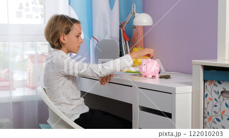 A young girl sits at her desk focused on placing coins into a piggy bank while writing notes about her savings. She learns the importance of managing personal finances 122010203