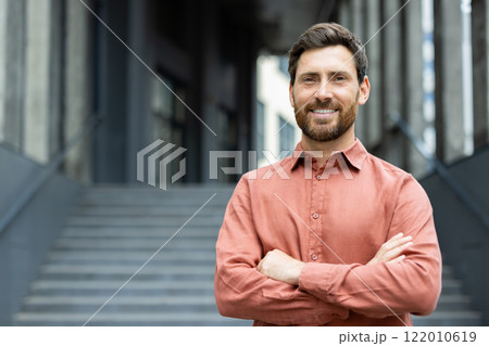 Portrait of confident and successful businessman outside office building. Man in red shirt smiling and looking at camera with arms crossed. 122010619