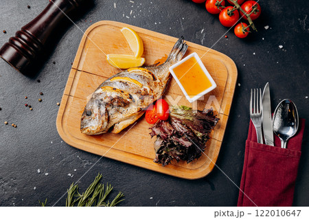 Grilled fish with lemon slices, salad, and dipping sauce on a wooden board for perfect seafood meal Grilled fish with lemon slices, salad, and dipping sauce on a wooden board for perfect seafood meal 122010647