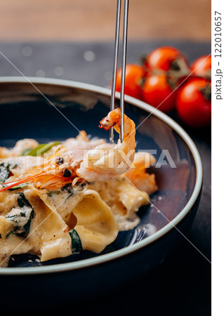 Close-up of a shrimp lifted with tongs from creamy pasta in a dark bowl 122010657