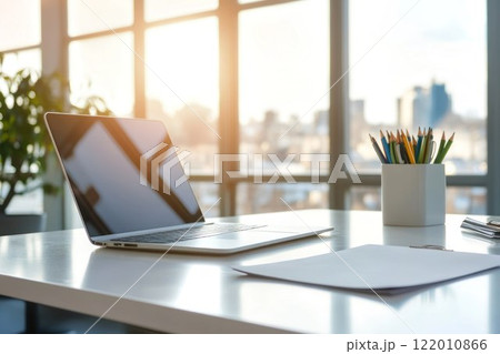 Laptop on a white table in a sleek office, nearby document paper and pencil holder, table positioned near a large window, bright sunlight. Laptop on a white table in a sleek office, nearby document paper and pencil holder, table positioned near a large window, bright sunlight. 122010866