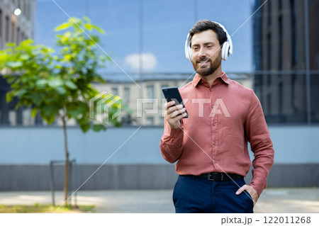 Successful businessman wearing headphones and with phone in hand walks through the business center of the city, office worker happily watches online video, dressed in red shirt. Successful businessman wearing headphones and with phone in hand walks through the business center of the city, office worker happily watches online video, dressed in red shirt. 122011268