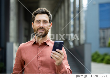 Portrait of successful businessman man with phone in hands. Office worker smiling and looking at camera, using app on smartphone, walking in city from outside office building. Portrait of successful businessman man with phone in hands. Office worker smiling and looking at camera, using app on smartphone, walking in city from outside office building. 122011274