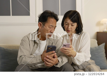夜のリビングで悩みながらスマホを使う熟年夫婦 122014264