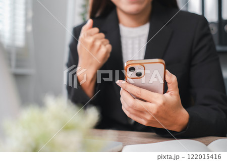 A professional woman in a suit celebrates a successful moment while looking at her smartphone in a contemporary office environment. 122015416