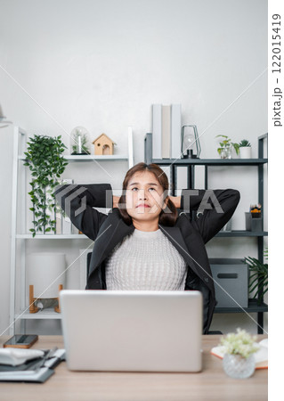 Businesswoman in a black suit relaxing at her desk with a laptop in a modern office setting with plants and shelves. Businesswoman in a black suit relaxing at her desk with a laptop in a modern office setting with plants and shelves. 122015419