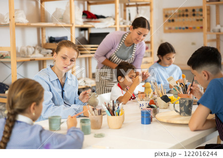 Children paint ceramic work-pieces at table with teacher in art studio 122016244