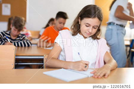 Interested preteen girl studying in classroom with classmates Interested preteen girl studying in classroom with classmates 122016701
