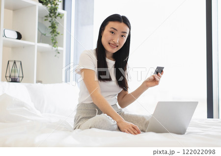 Asian woman sits on bed uses laptop for online shopping holding credit card. Casual home setting. Mid 20s enjoying convenience of internet shopping. Asian woman sits on bed uses laptop for online shopping holding credit card. Casual home setting. Mid 20s enjoying convenience of internet shopping. 122022098