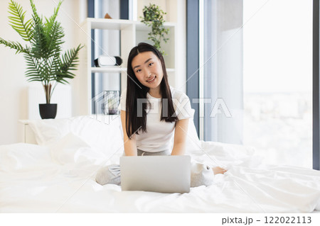 Young Asian woman sitting on bed using laptop in bright modern bedroom. She is smiling, creating relaxed and focused atmosphere, representing education and technology. 122022113