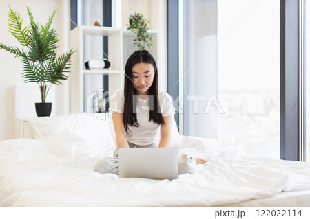 Asian female in early twenties works on laptop in bright bedroom setting. Casual and relaxed atmosphere emphasizes work-life balance. Asian female in early twenties works on laptop in bright bedroom setting. Casual and relaxed atmosphere emphasizes work-life balance. 122022114