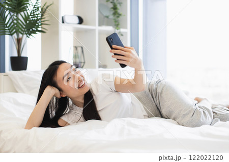 Young Asian woman having online video call from comfortable bed, smiling joyfully. Casual and relaxed setting with smartphone. Bright and airy room, expressing happiness and connection. Young Asian woman having online video call from comfortable bed, smiling joyfully. Casual and relaxed setting with smartphone. Bright and airy room, expressing happiness and connection. 122022120