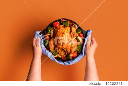 Roasted chicken in an iron cast, with vegetables, minimalist on an orange background. Roasted chicken in an iron cast, with vegetables, minimalist on an orange background. 122022414