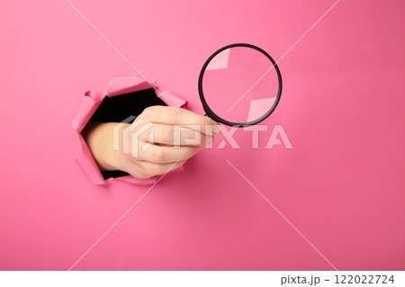 Female hand with magnifying glass punching through the pink paper. Female hand with magnifying glass punching through the pink paper. 122022724