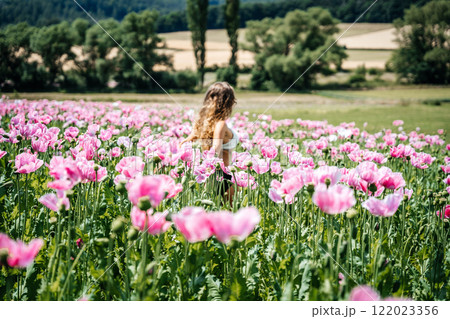 A girl with long, curly blonde hair stands in a field of pink poppies, surrounded by a stunning mountain landscape. The flowers sway gently in the wind, creating a serene and picturesque scene 122023356