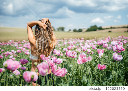 A girl with long, curly blonde hair stands in a field of pink poppies, surrounded by a stunning mountain landscape. The flowers sway gently in the wind, creating a serene and picturesque scene 122023360