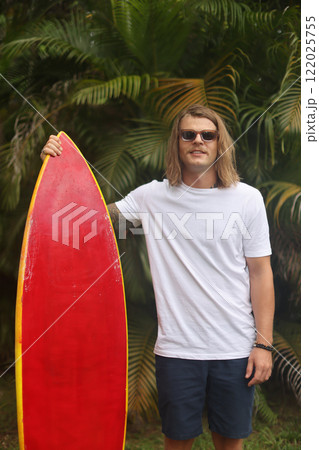A surfer is depicted riding a red board amidst a vibrant tropical setting, basking in the sun 122025755