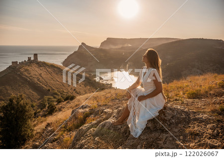 A woman in a white dress sits on a rock overlooking a body of water. The scene is serene and peaceful, with the woman enjoying the view and the calmness of the surroundings. 122026067