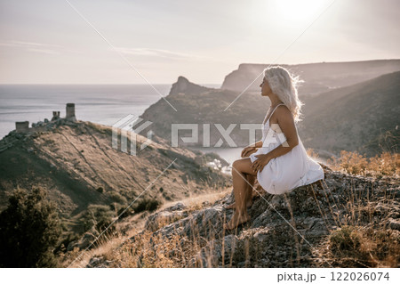 A woman is sitting on a hillside overlooking the ocean. She is wearing a white dress and has blonde hair. The scene is serene and peaceful, with the ocean in the background. 122026074