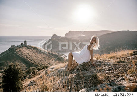 A woman is sitting on a hillside overlooking the ocean. She is wearing a white dress and has blonde hair. The scene is serene and peaceful, with the ocean in the background. 122026077
