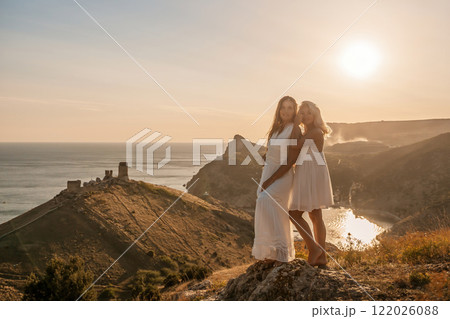 Two young girls are standing on a hillside, one of them wearing a white dress. The sun is shining brightly, creating a warm and inviting atmosphere. The girls seem to be enjoying their time together. Two young girls are standing on a hillside, one of them wearing a white dress. The sun is shining brightly, creating a warm and inviting atmosphere. The girls seem to be enjoying their time together. 122026088