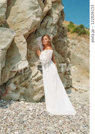 A woman in a white dress is standing on a rocky beach. Concept of serenity and tranquility, as the woman is enjoying the natural beauty of the beach. A woman in a white dress is standing on a rocky beach. Concept of serenity and tranquility, as the woman is enjoying the natural beauty of the beach. 122026155