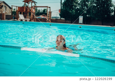 A woman is swimming in a pool. She is wearing a green top and is smiling. The pool is blue and has a few chairs around it. 122026199