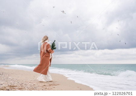 Blond woman Christmas tree sea. Christmas portrait of a happy woman walking along the beach and holding a Christmas tree on her shoulder. She is wearing a brown coat and a white suit. 122026701