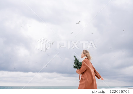 Blond woman Christmas tree sea. Christmas portrait of a happy woman walking along the beach and holding a Christmas tree on her shoulder. She is wearing a brown coat and a white suit. 122026702