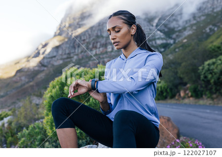 Young sportswoman checking pulse while sitting on a rock in a natural park. Female looking at smartwatch while sitting outdoors in a natural park.  122027107