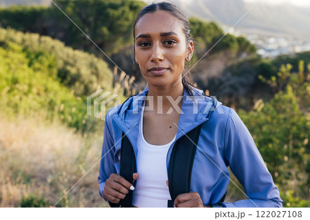 Portrait of a beautiful sportswoman wearing a backpack and looking at camera while standing in a natural park at sunset  122027108