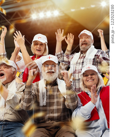 Enthusiastic group of elderly people, fans with hands raised, celebrating in brightly lit stadium during thrilling sports match 122028280