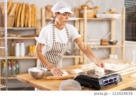 Female baker prepares raw dough, cuts pieces and weighs them on scale to make buns or croissants Female baker prepares raw dough, cuts pieces and weighs them on scale to make buns or croissants 122028638